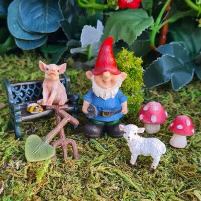 A garden gnome figurine with a red hat and blue shirt standing next to a small white lamb figurine, with a pink pig figurine sitting on a black bench behind. In the foreground, there's a miniature bee on a brown fence, and two red-capped mushroom figurines are nearby. The setting looks like a lush garden with mossy ground cover and green plants in the background.