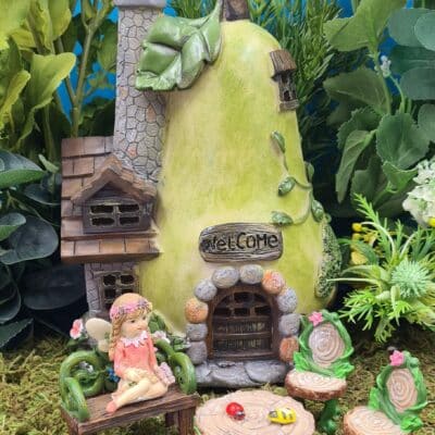 A whimsical fairy garden scene featuring a fairy sitting on a bench with a pear-shaped house adorned with a "Welcome" sign, a stone chimney, windows, and leaf accents. In front are a table and chairs shaped like tree stumps with tiny insect decorations, all set against a backdrop of assorted artificial plants and a bright blue sky.