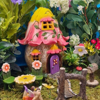 Decorative fairy garden featuring a whimsical house made from a coconut with a pink blossom roof, yellow highlights, and a purple door, surrounded by artificial plants, flowers, and a fairy figure.