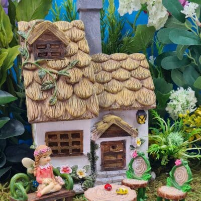 A miniature fairy garden scene featuring a detailed cottage with a straw roof, surrounded by artificial plants and flowers with a figurine of a fairy seated on a bench.