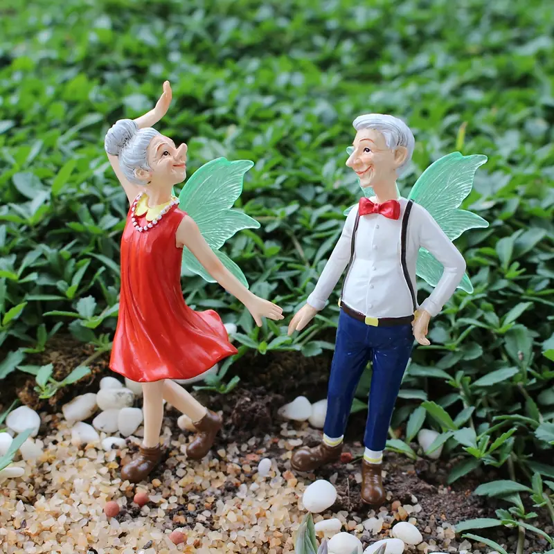 Two whimsical fairy figurines, an elderly woman in a red dress and an elderly man in a white shirt and bowtie, joyfully dancing together amidst lush greenery.