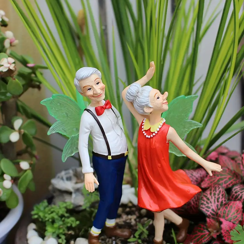 Charming garden figurines of an elderly couple, with the man in a white shirt and red bow tie and the woman in a red dress, both adorned with colorful wings, surrounded by lush greenery.