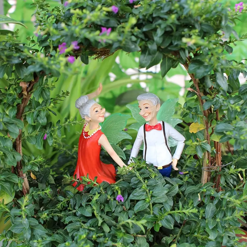 A playful scene featuring two fairy figurines, an elderly couple, with green wings, surrounded by lush greenery and colorful flowers, highlighting a cheerful interaction between them.