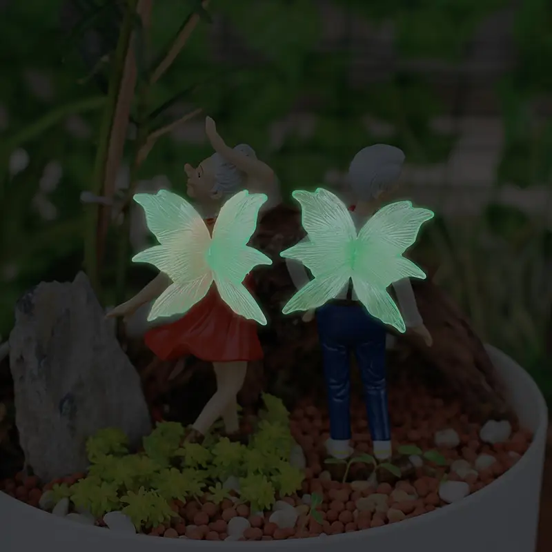Two whimsical figurines with glowing butterfly wings standing in a garden planter surrounded by small plants and pebbles.