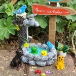 A whimsical garden scene featuring a miniature gnome workshop sign, a stone fountain with colorful frogs and ducks, and playful animal figurines including dogs and ladybugs, surrounded by lush green foliage.