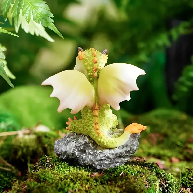 A cute dragon figurine with yellow wings perched on a rock, surrounded by lush greenery and moss.