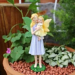 A delicate fairy figurine depicting a mother with wings holding her child, surrounded by lush greenery and decorative stones in a flowerpot.