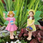 Two whimsical fairy figurines in colorful dresses, one holding a book and the other a basket of fruit, surrounded by lush greenery and decorative stones.