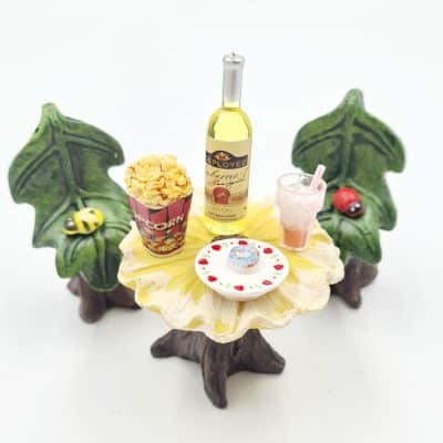 A whimsical table featuring miniature popcorn, a bottle of wine, a pink drink with a straw, and a decorated dessert, set among leaf-shaped chairs.