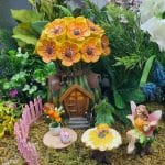 A whimsical miniature fairy garden featuring a flower-adorned cottage, adorable decorations, and a fairy sitting at a mushroom table.