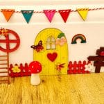A colorful handcrafted fairy house scene featuring a yellow door, decorative accents like flowers, a rainbow, and small figurines, set against a wooden background.