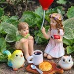 A charming scene featuring fairy figurines enjoying a tea party with a bluebird, owl, and ladybug, surrounded by lush greenery.