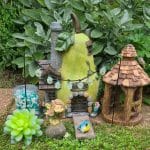 A vibrant fairy garden featuring a pear-shaped house, a small gazebo, decorative stones, a figure of a fairy, a succulent plant, and a colorful bird, all surrounded by lush green foliage.