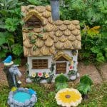 A whimsical miniature cottage with a thatched roof, surrounded by vibrant foliage, featuring a small fountain and colorful garden ornaments.